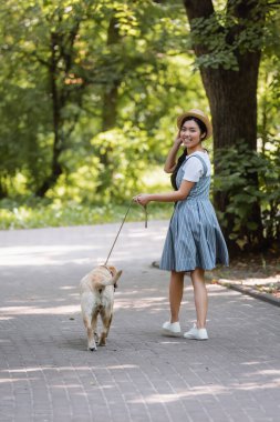 Pozitif Asyalı kadın çizgili elbisesiyle parkta köpekle yürüyor ve kameraya gülümsüyor.