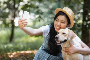 Hasır şapkalı neşeli Asyalı kadın parkta sarı labradorla selfie çekiyor.