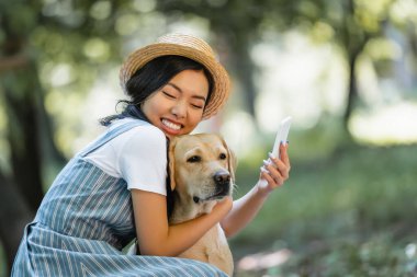 Gözleri kapalı mutlu Asyalı kadın parkta selfie çekerken Labrador 'a sarılıyor.