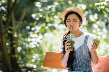 Gözleri kapalı Asyalı kadın parkta bankta taze smoothie içiyor.