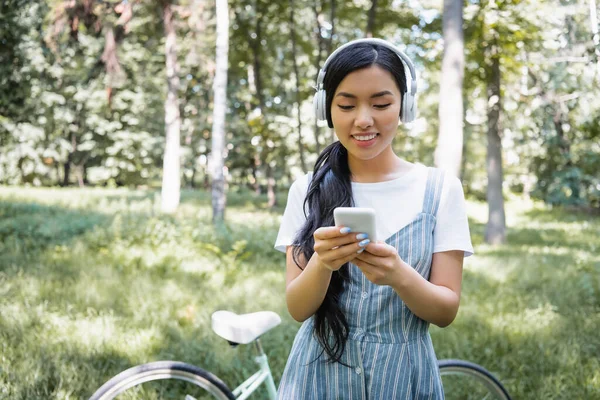 Bulanık Bisikletin Yanında Cep Telefonu Kullanan Kulaklıklı Mutlu Asyalı Kadın — Stok fotoğraf
