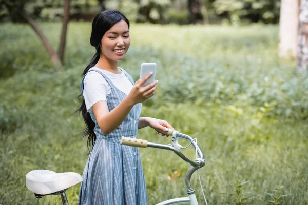 穿着条纹太阳衣的快乐的亚洲女人看着自行车旁边的手机 — 图库照片