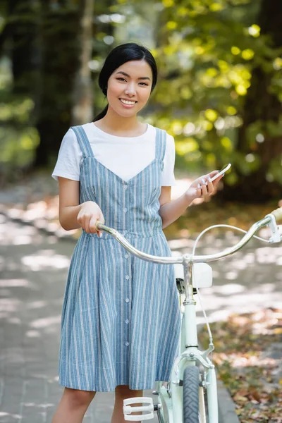 Parkta Bisiklete Gülümseyen Akıllı Telefonlu Genç Asyalı Kadın — Stok fotoğraf