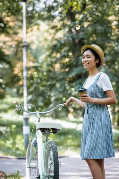 紙コップを持ったアジア系の女性が公園で自転車の近くで笑っています — ストック写真