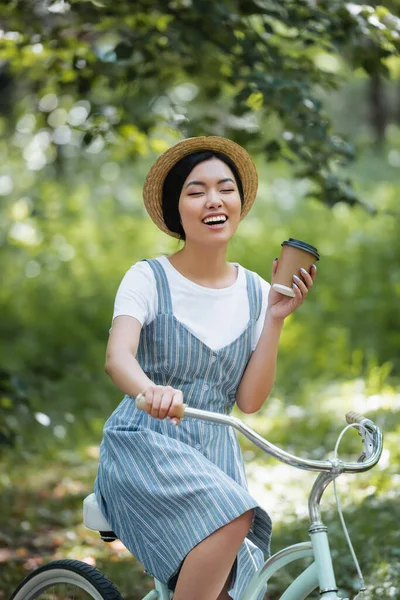 一个快乐的亚洲女人 闭着眼睛笑着 一边骑自行车 一边喝着外卖的饮料 — 图库照片