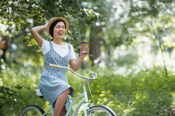 コーヒーを飲み公園で目を閉じたまま自転車で笑う陽気なアジアの女性 — ストック写真