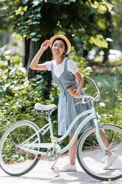 公園のバイクの近くのカメラで笑顔を見せながら麦藁帽子を調整する若いアジア人女性 — ストック写真