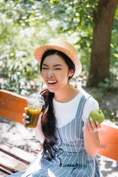 Vzrušená Asijská Žena Slamáku Drží Čerstvé Smoothie Zralé Jablko Lavičce — Stock fotografie