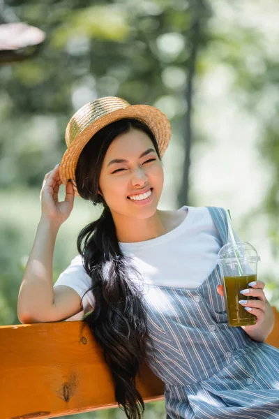 Glückliche Asiatin Mit Plastikbecher Smoothie Berührt Strohhut Auf Bank Park — Stockfoto