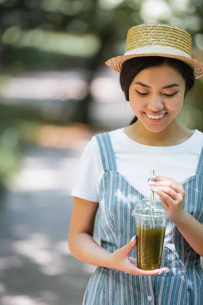 快乐的亚洲女人 戴着草帽 在户外端着塑料杯子 — 图库照片
