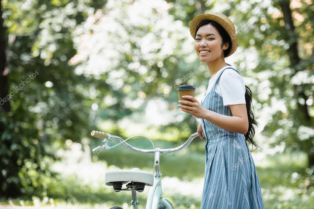 happy asian woman with takeaway drink looking at camera near bicycle in park