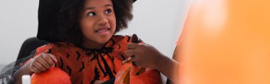happy african american girl in witch costume looking at brother carving pumpkin, banner clipart