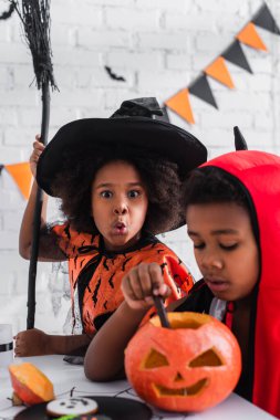 blurred african american boy in halloween costume carving pumpkin near spooky sister with broom  clipart