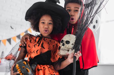 spooky african american kids in halloween costumes holding skull, carved pumpkin and broom  clipart