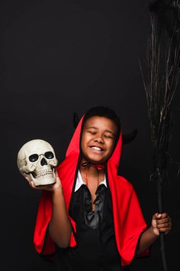 happy african american boy holding broom and skull isolated on black  clipart