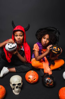 spooky african american children in halloween costumes holding skull and broom near pumpkins on black  clipart