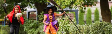 cheerful african american kids in halloween costumes holding brooms outside, banner clipart