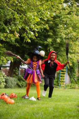 Cadılar Bayramı kostümlü neşeli Afro-Amerikalı çocuklar ellerinde süpürgelerle arka bahçede 