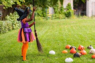 african american kid in witch halloween costume holding broom near carved pumpkins on lawn  clipart