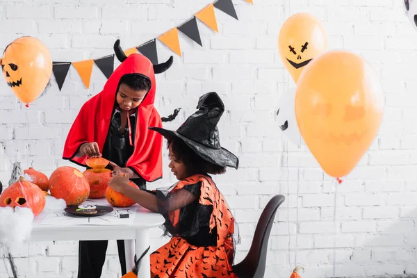 Crianças Americanas Africanas Sorridentes Trajes Halloween Esculpindo Abóboras Perto Biscoitos — Fotografia de Stock