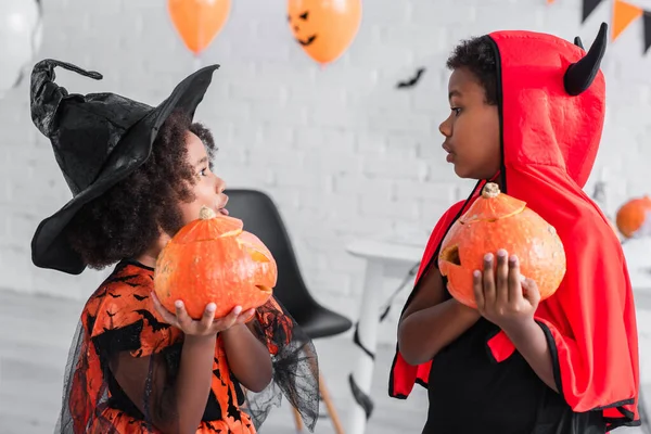 side view of surprised african american kids in halloween costumes holding carved pumpkins at home
