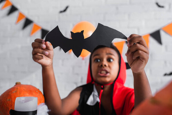 blurred and scared african american boy in halloween costume holding paper cut bat
