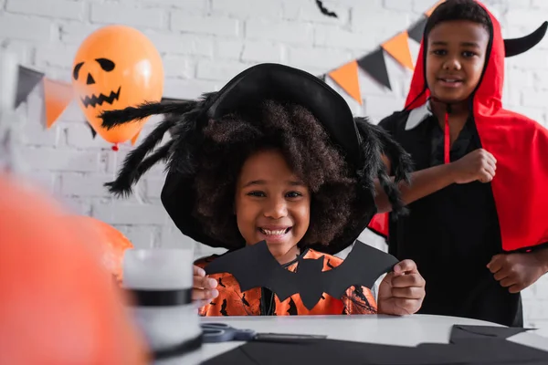 happy african american girl in halloween costume holding paper cut bat near brother