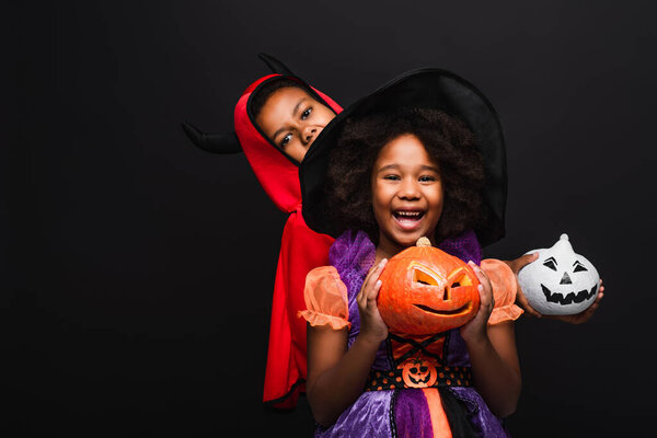 cheerful african american kids in halloween costumes holding spooky pumpkins isolated on black 