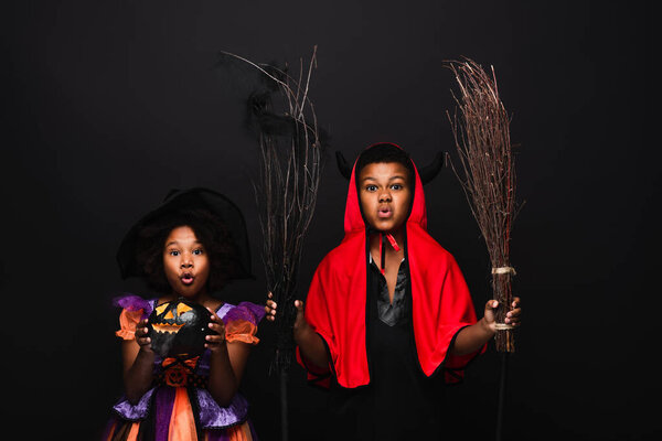 african american girl in halloween costume holding carved pumpkins near brother with brooms isolated on black 