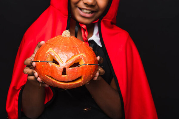 cropped view of happy african american boy in devil halloween costume holding carved pumpkin isolated on black