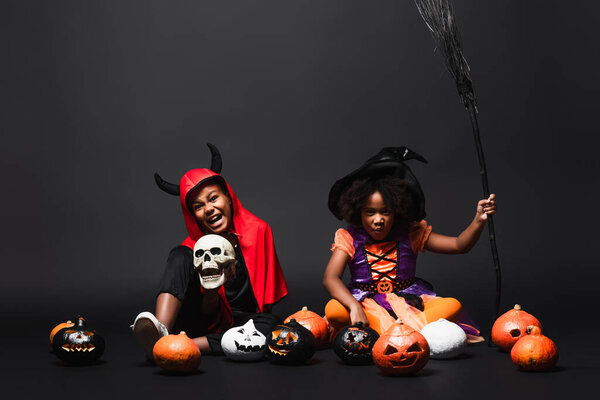 african american children in halloween costumes holding skull and broom near pumpkins on black 