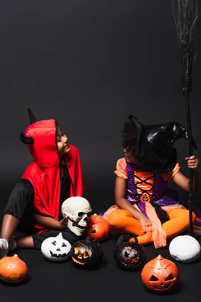 african american kids in halloween costumes holding skull and broom near pumpkins on black 