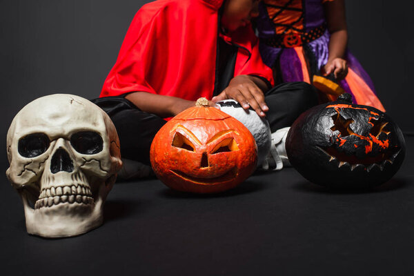 cropped view of african american siblings in halloween costumes holding carved pumpkins on black 