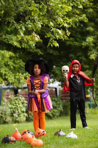 african american kids in halloween costumes holding brooms on backyard 