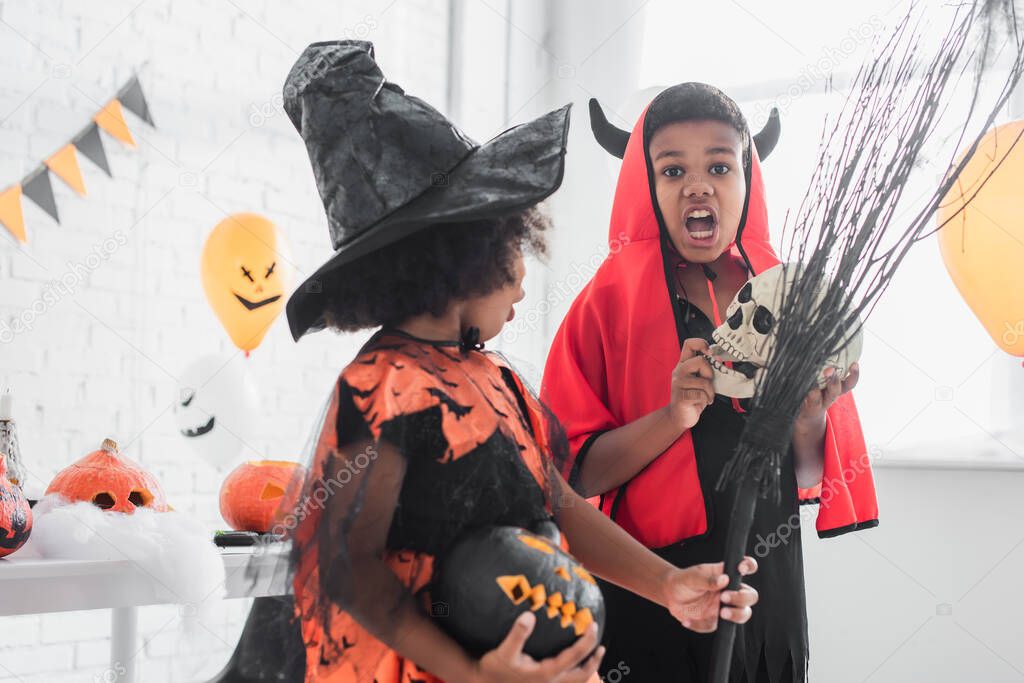 spooky african american boy in halloween costume holding skull near sister with broom and carved pumpkin 
