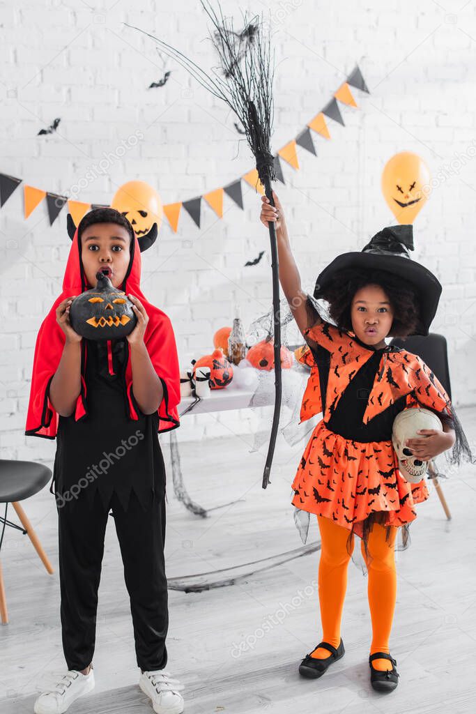 spooky african american girl in halloween costume holding skull and broom near brother with carved pumpkin 