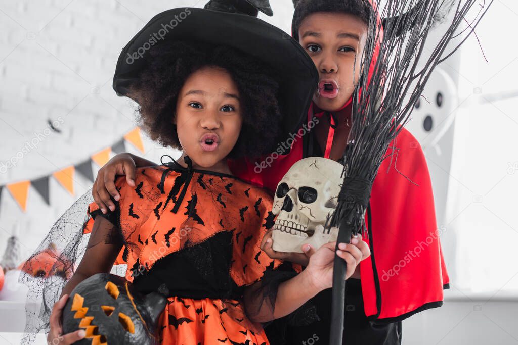 spooky african american kids in halloween costumes holding skull, carved pumpkin and broom 