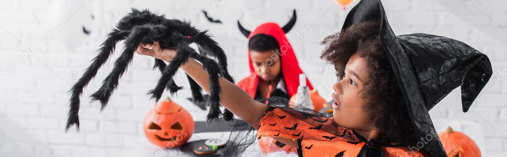 african american girl in witch costume holding toy spider near blurred brother, banner