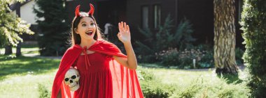 amazed girl in devil halloween costume holding spooky skull and waving hand, banner clipart