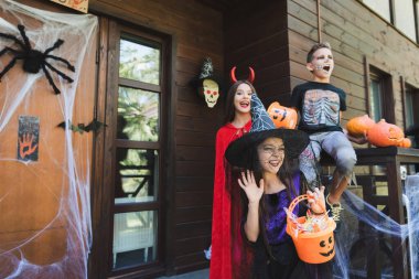 Cadı şapkalı kız verandada süslenmiş ürkütücü cadılar bayramı kostümlü heyecanlı çocukların yanında el sallıyor.