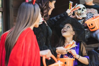 Cadılar bayramı kostümü giymiş bir kadın elinde kovalı çocukların yanında şeker tutuyor.