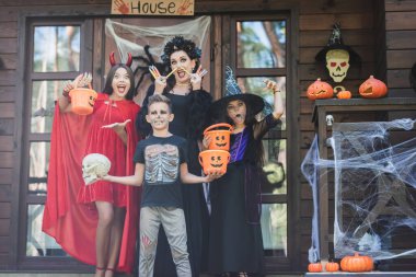 Cadılar Bayramı kostümlü çocuklar verandada ellerinde kovalarla, şekerlerle ve kafataslarıyla duruyorlar.