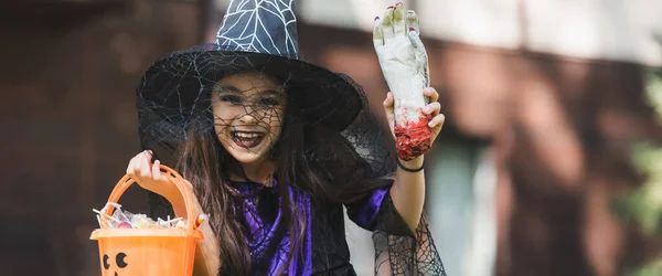 cheerful girl in witch hat holding bucket with sweets and toy hand, banner