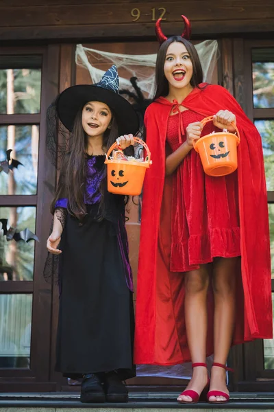 Užaslé Sestry Čarodějnických Ďábelských Halloweenských Kostýmech Kbelíky Sladkostí Verandě Chalupy — Stock fotografie