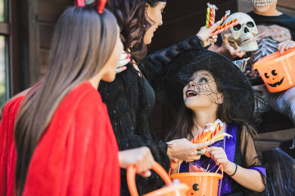 woman in halloween costume holding candies near amazed kids with buckets