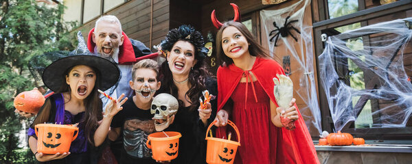 creepy family in halloween costumes grimacing while holding buckets and sweets near house, banner