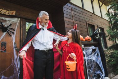 Mutlu kızın yanında, verandada şeytan kostümü giymiş heyecanlı bir adam var.