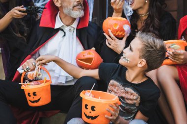 Cadılar bayramı kostümü giymiş, ailesinin yanındaki kovadan şeker alan çocuk.