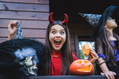 užaslá dívka v ďábel kostým v blízkosti sestra v čarodějnice klobouk a vyřezávané dýně na chalupě veranda