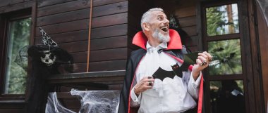 laughing middle aged man in halloween vampire costume holding paper cut bat on porch of wooden house, banner clipart